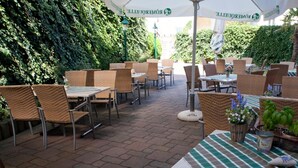 Un espacio al aire libre con patio, mobiliario de mimbre, mesas y sombrillas, rodeado de vegetación.