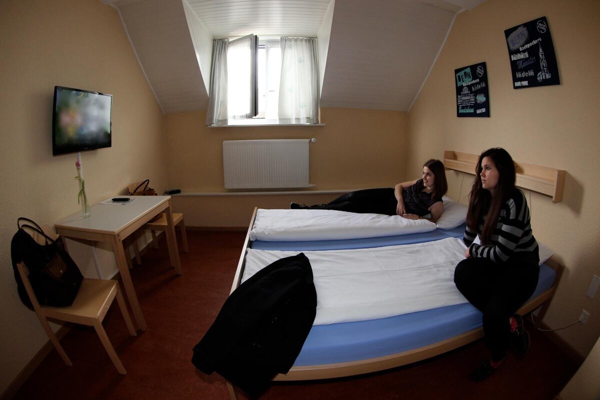 A dormitory room with two beds, a desk, a chair, a TV, and a window with curtains.