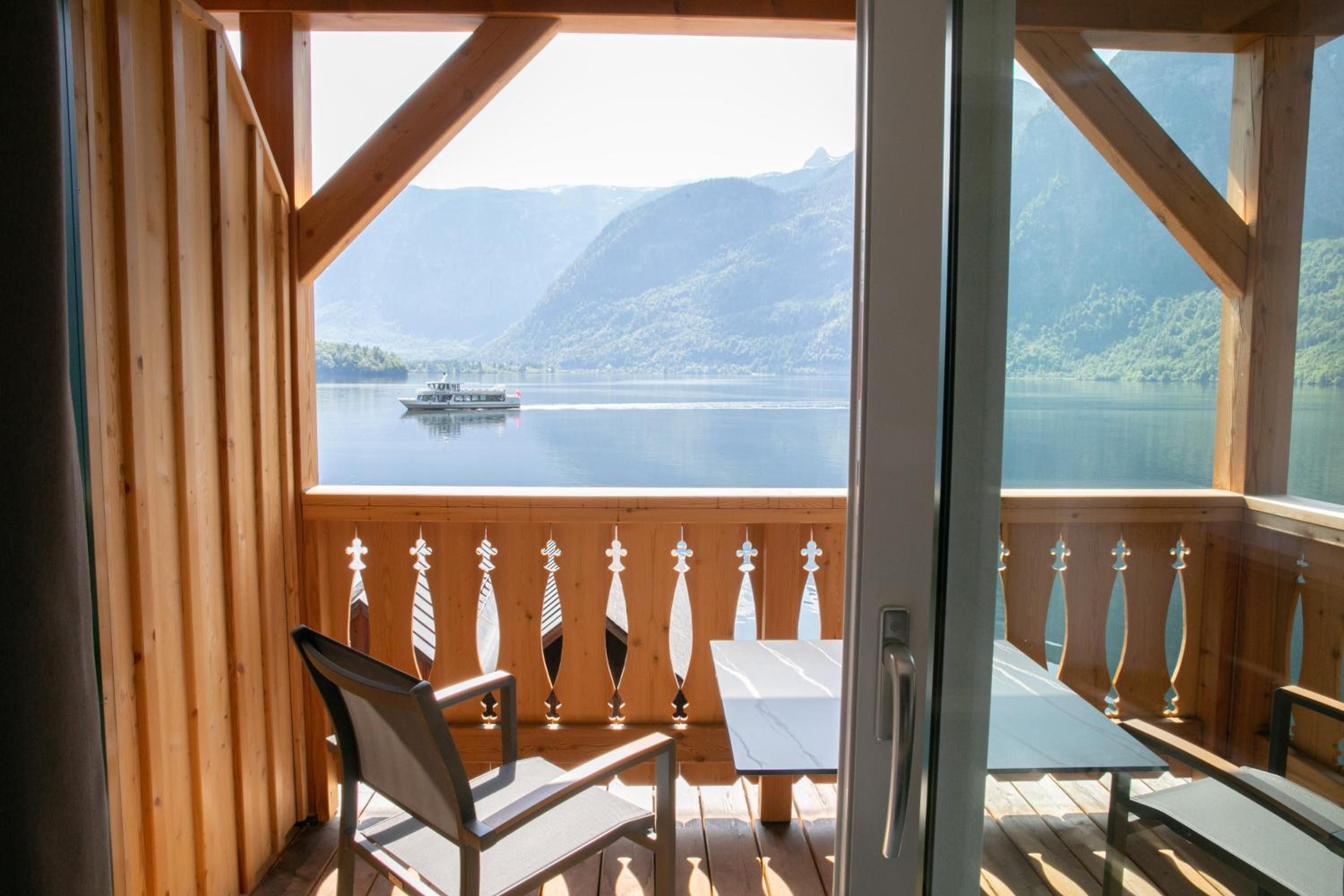 A wooden deck with a table and chairs, overlooking a calm lake with a boat and mountains in the distance.