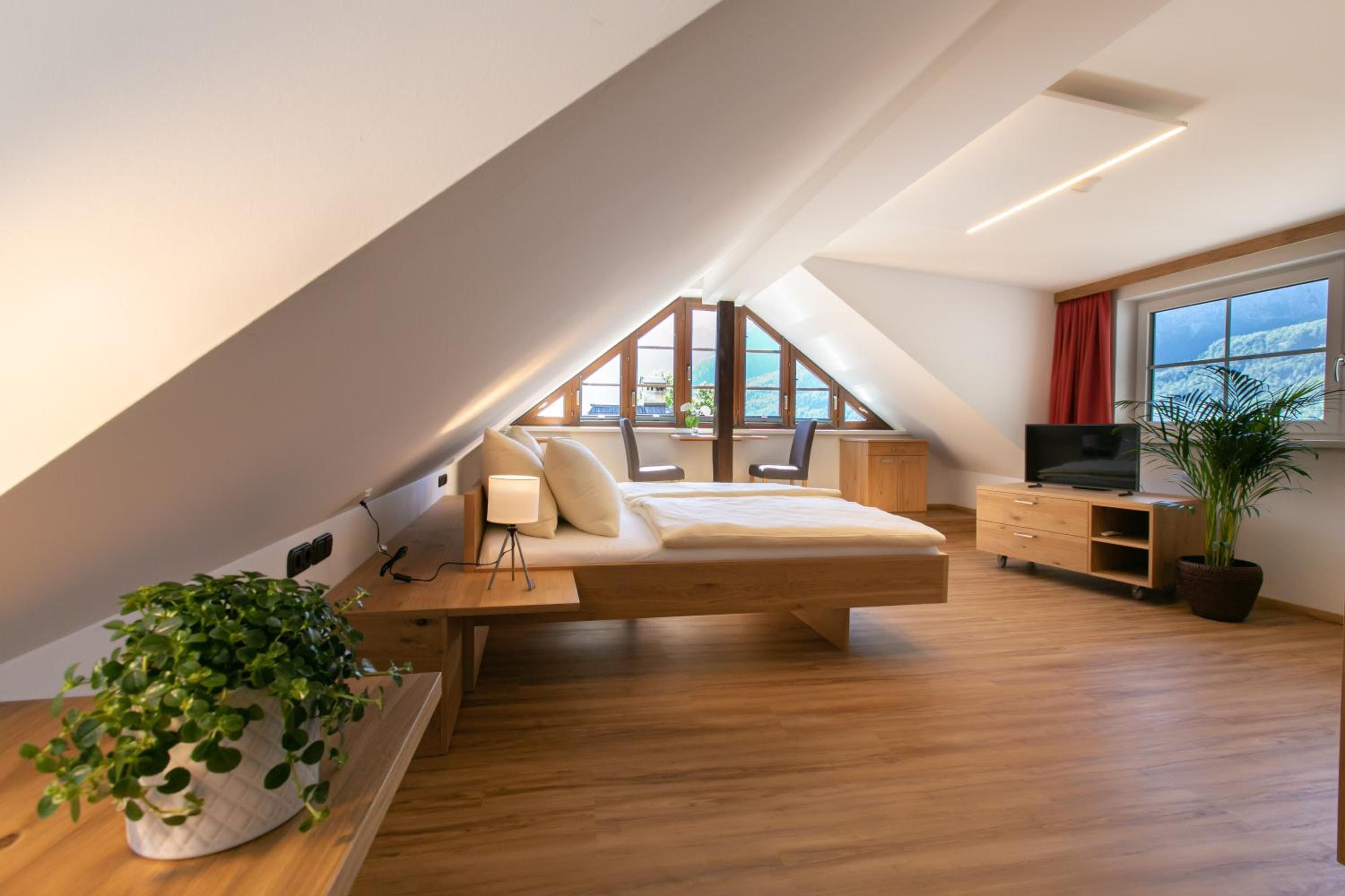 Un dormitorio estilo loft moderno con techo inclinado, cama, escritorio, televisor y un ventanal con vistas.