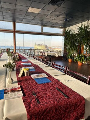 Una larga mesa de comedor con manteles rojos y sillas, con un paisaje urbano y una masa de agua como fondo.