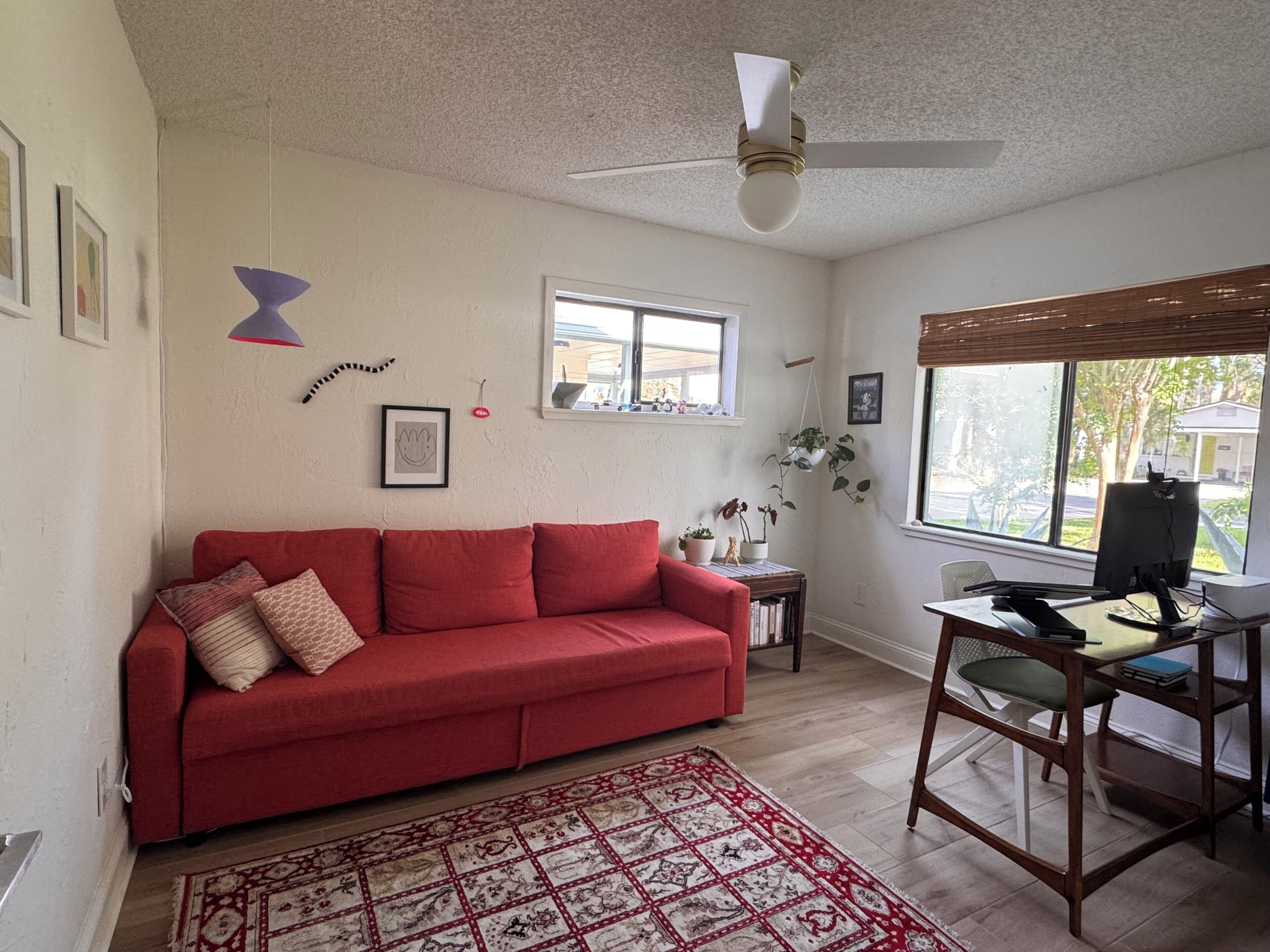 A room with a red sofa, a desk with a computer, a ceiling fan, a window, and a patterned rug.
