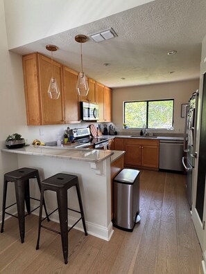 A kitchen with wooden cabinets, a stainless steel refrigerator, a microwave, and a window with a view of trees. - Spacious, family-friendly beach house (Atlantic Beach)