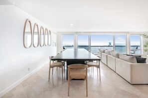 Ein modernes Interieur mit einem Esstisch, Stühlen, einer Couch und großen Fenstern mit Blick auf das Meer.
