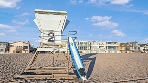 Une tour de surveillance, avec une planche de surf bleue appuyée contre, se dresse sur une plage de sable, avec des bâtiments visibles à l’arrière-plan.