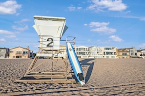 Ein Rettungsschwimmturm mit einem blauen Surfbrett, das dagegen gelehnt ist, auf einem Sandstrand, im Hintergrund sind Gebäude zu sehen.