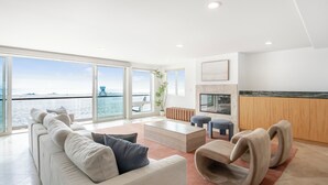 Un salon moderne doté d’une grande fenêtre donnant sur la mer, d’une cheminée et d’un coin cuisine avec des meubles en bois.
