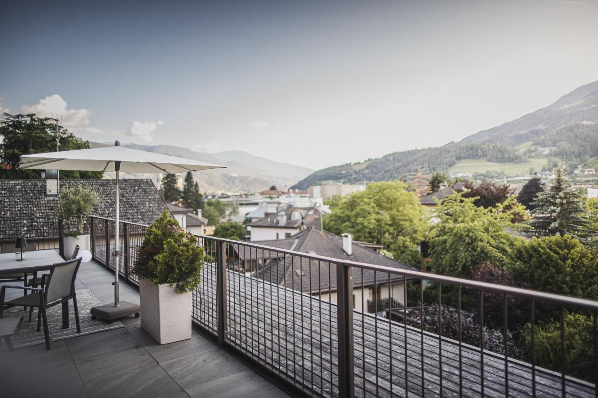 A terrace with a table and chairs, a potted plant, and a view of a town and mountains.