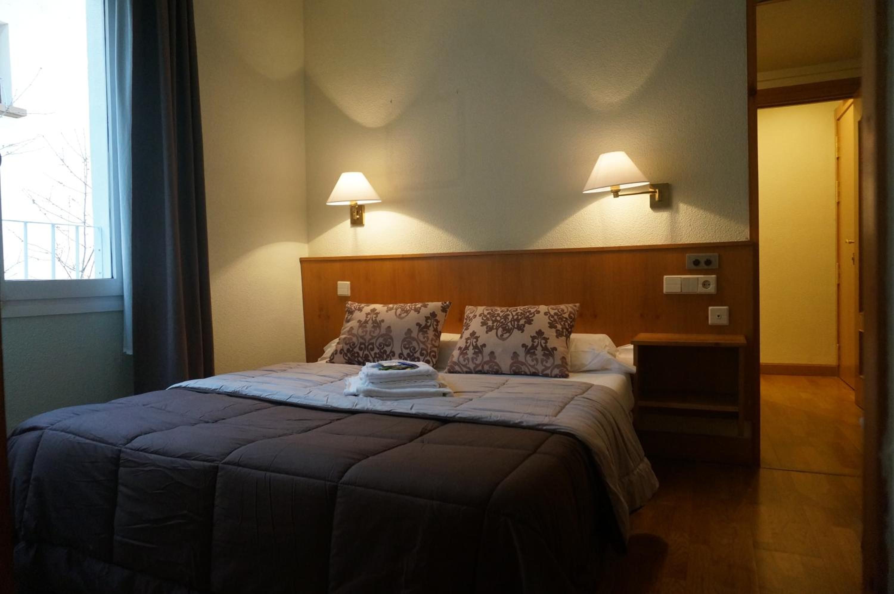 Habitación de hotel con cama, dos lámparas de pared, una mesita de noche y una ventana.