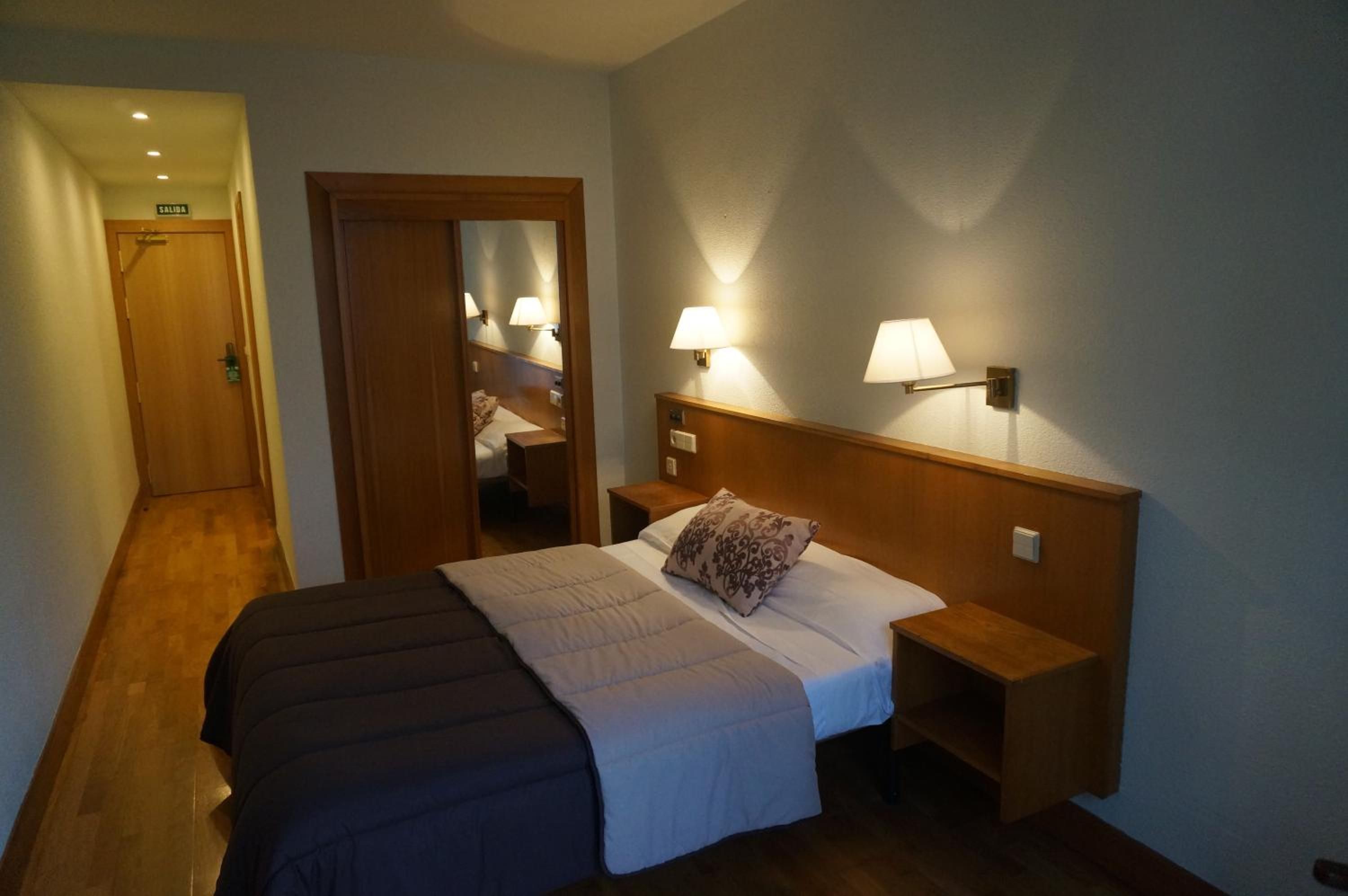 Habitación de hotel con cama, lámparas de noche, cabecero de madera y una puerta con la leyenda 'SALIDA'.