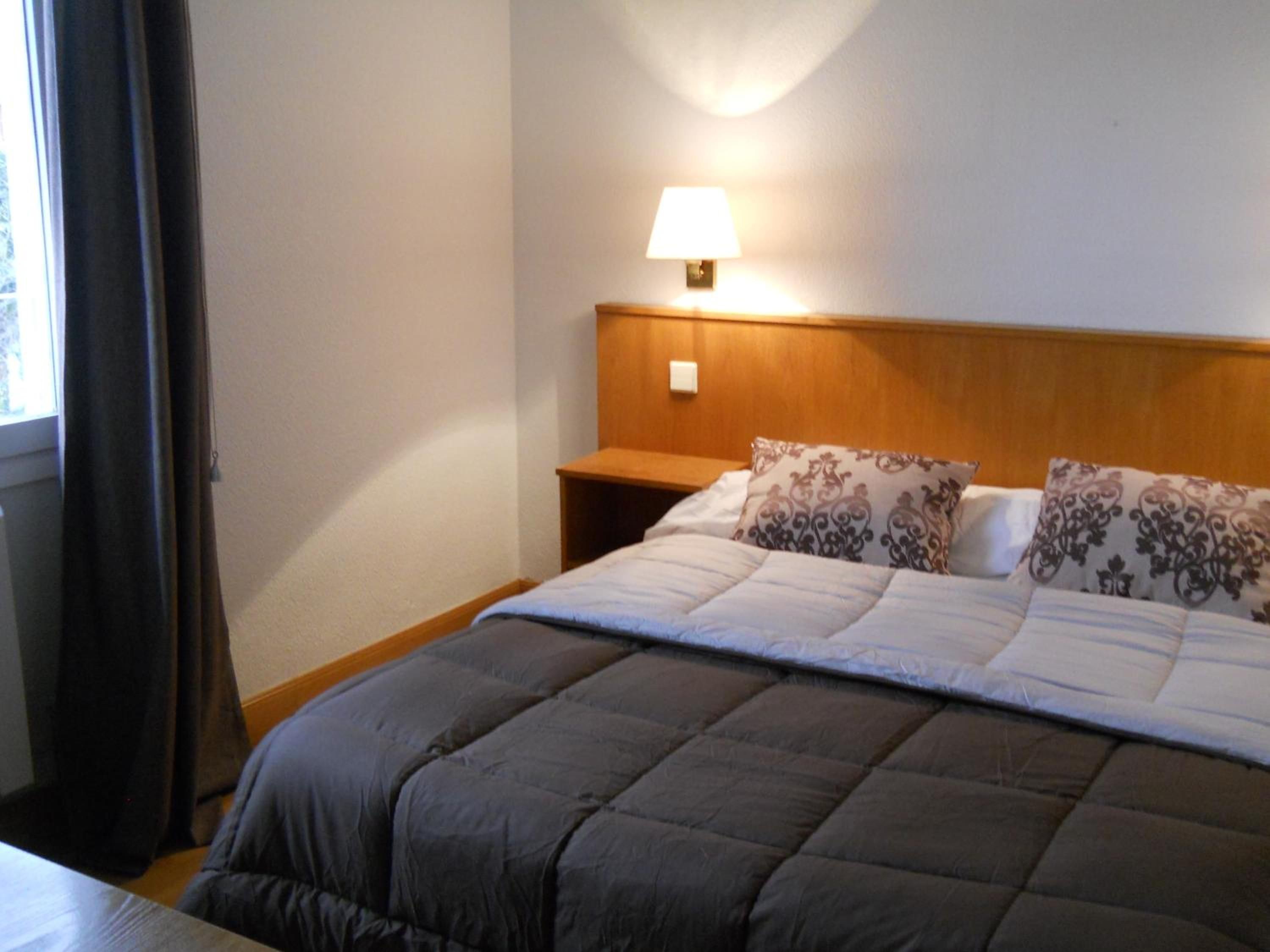 Habitación de hotel con cama, mesita de noche, lámpara y ventana con cortinas.
