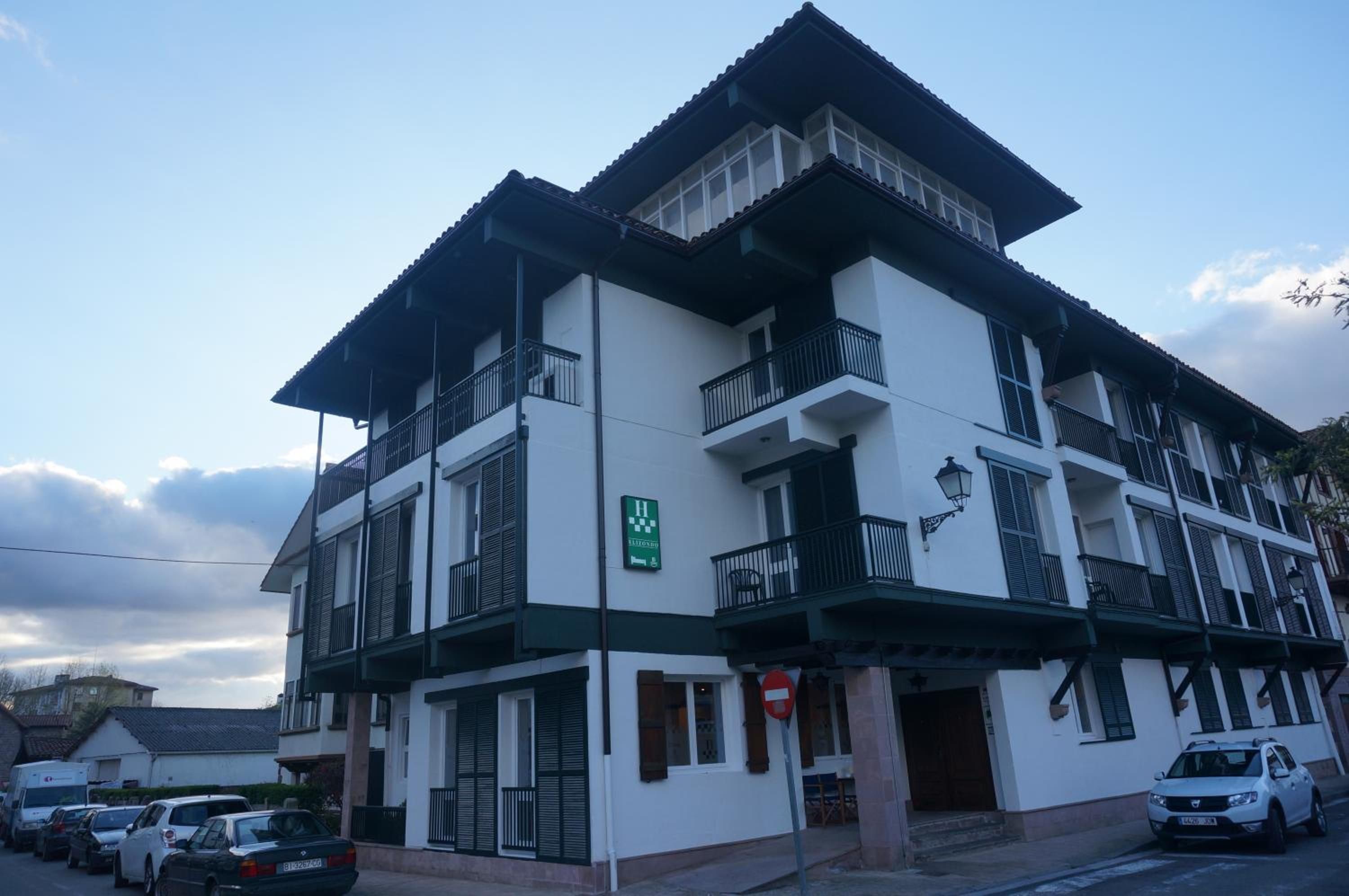 Un edificio de varios pisos con balcones, una señal que indica un hotel a la vista, y coches estacionados al frente.