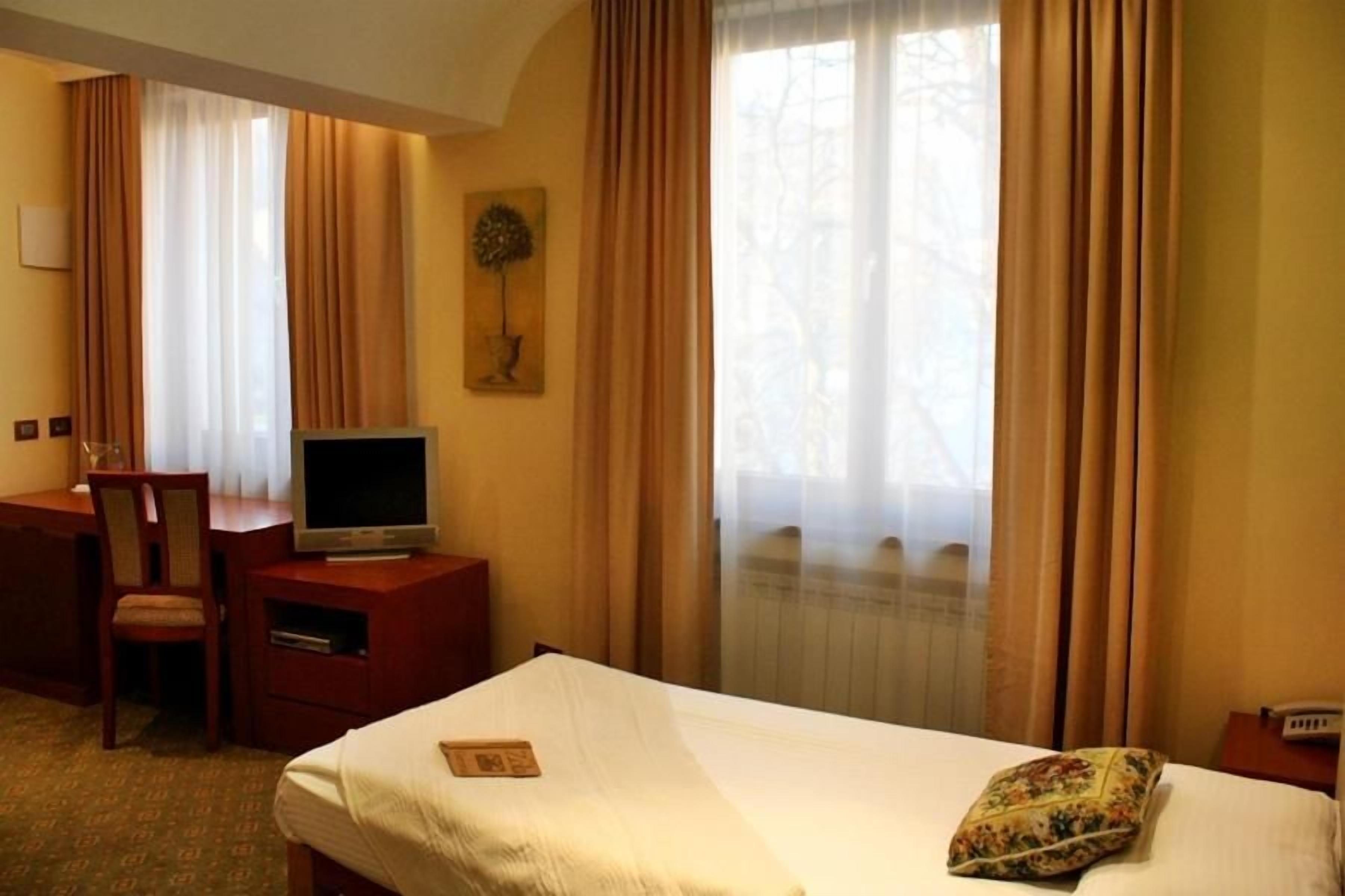 Habitación de hotel con cama, escritorio con televisor, una silla y ventana con cortinas.