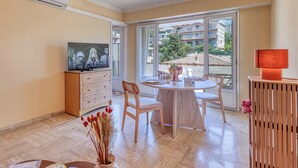 A modern living room with a television, a dining table, chairs, a wooden cabinet, and a large window with a view of buildings and trees. - Lion Apt - Near City Center - Terrace, Nice, France (Nice)