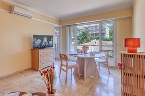 A modern living room with a television, a dining table, chairs, a wooden cabinet, and a large window with a view of buildings and trees. - Lion Apt - Near City Center - Terrace, Nice, France (Nice)