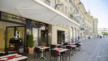 An outdoor cafe with tables and chairs, a gelateria across the street, and a building with a balcony.