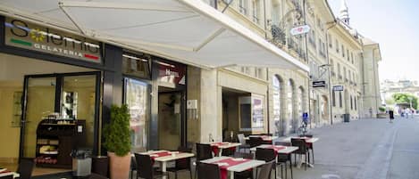An outdoor cafe with tables and chairs, a gelateria across the street, and a building with a balcony.