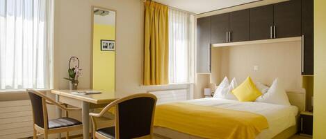 Habitación de hotel con una cama, un escritorio, dos sillas, un espejo y una ventana con cortinas.
