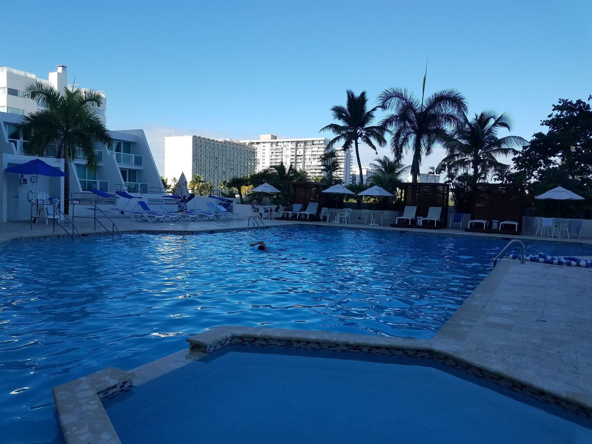 Piscina con sillones de descanso y sombrillas, rodeada de palmeras y edificios modernos.