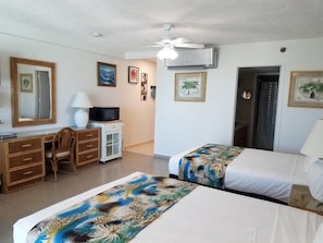 Habitación de hotel con dos camas, un escritorio con silla, microondas, ventilador de techo y cuadros en las paredes.