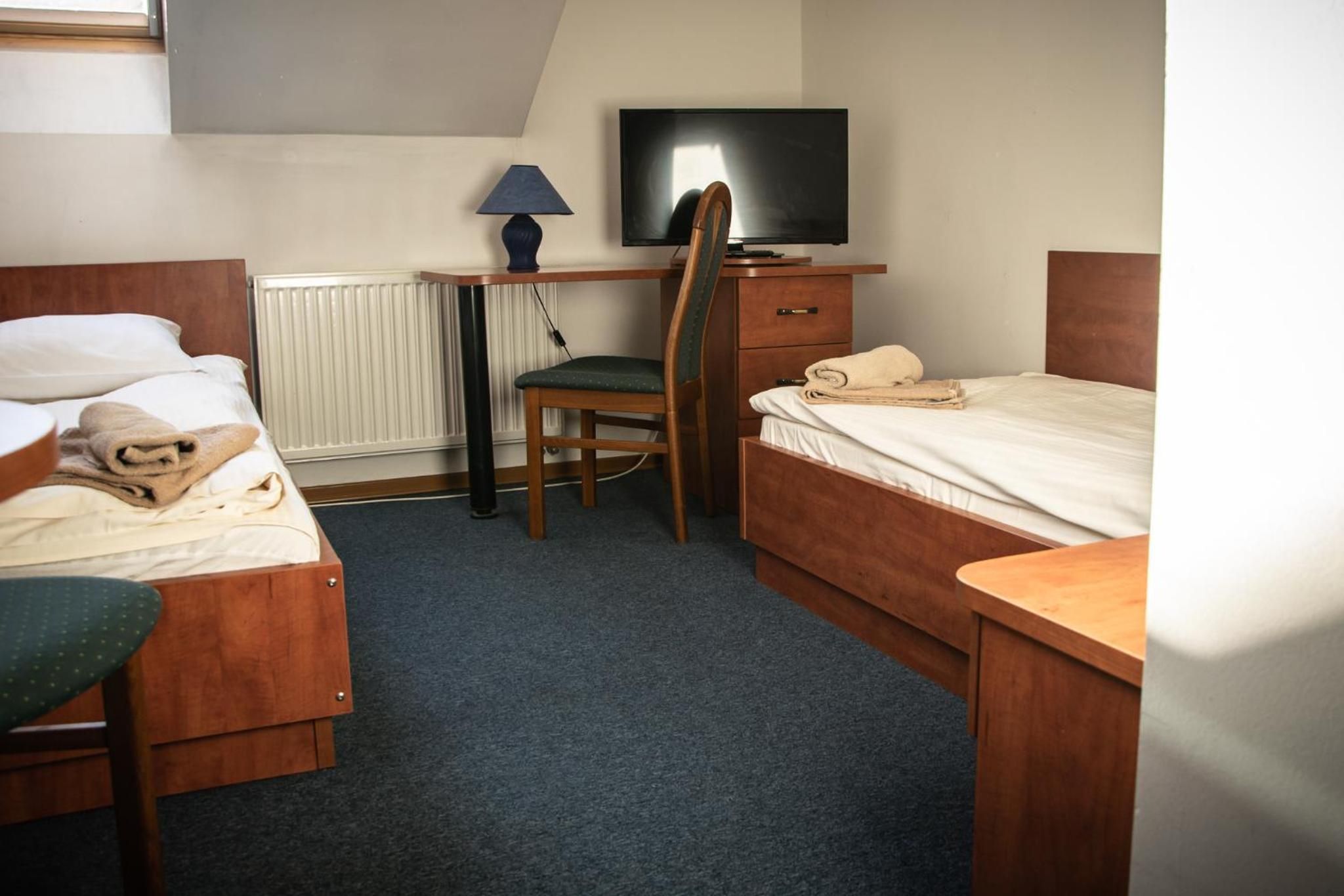 A hotel room with two single beds, a desk with a chair, a television, and a radiator.