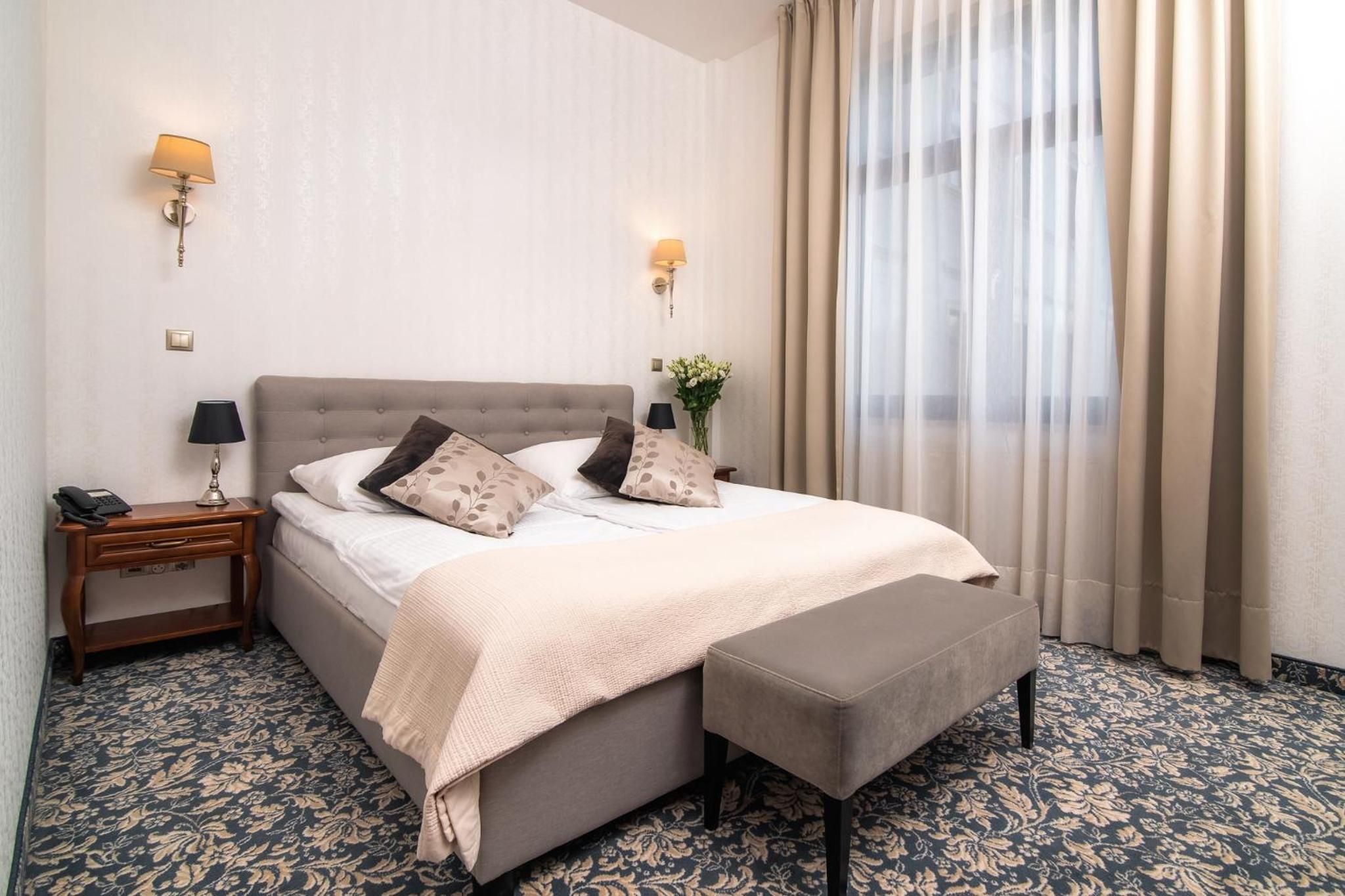 Ein Hotelzimmer mit einem Bett, Nachttischen, Lampen, einem Fenster mit Vorhängen und einem gemusterten Teppich.