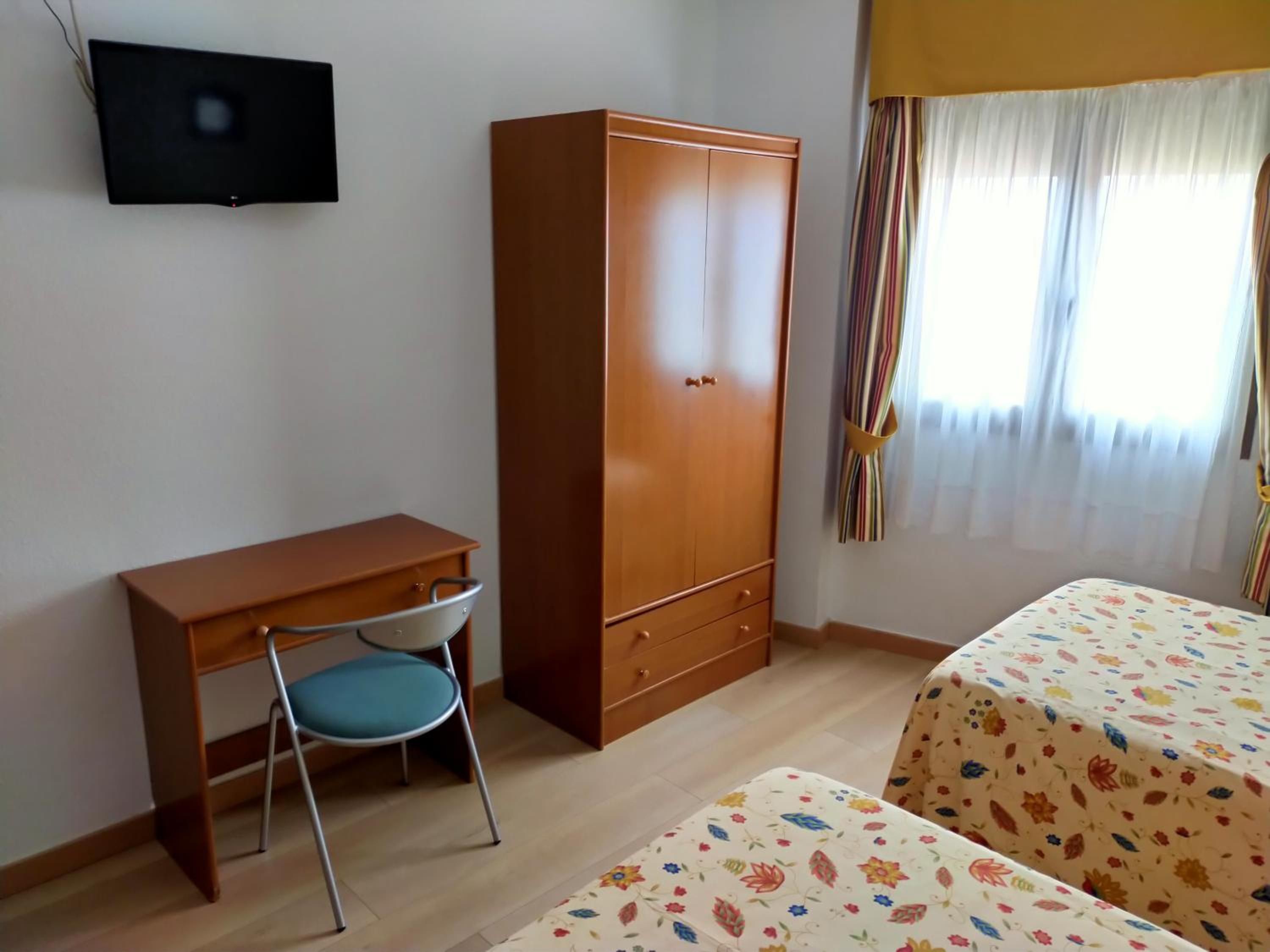 A room with a wooden desk, a chair, a wardrobe, and two beds with floral bedding.