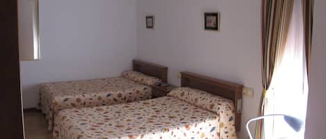 A small, simple room with two beds, a chair, and a window with curtains.