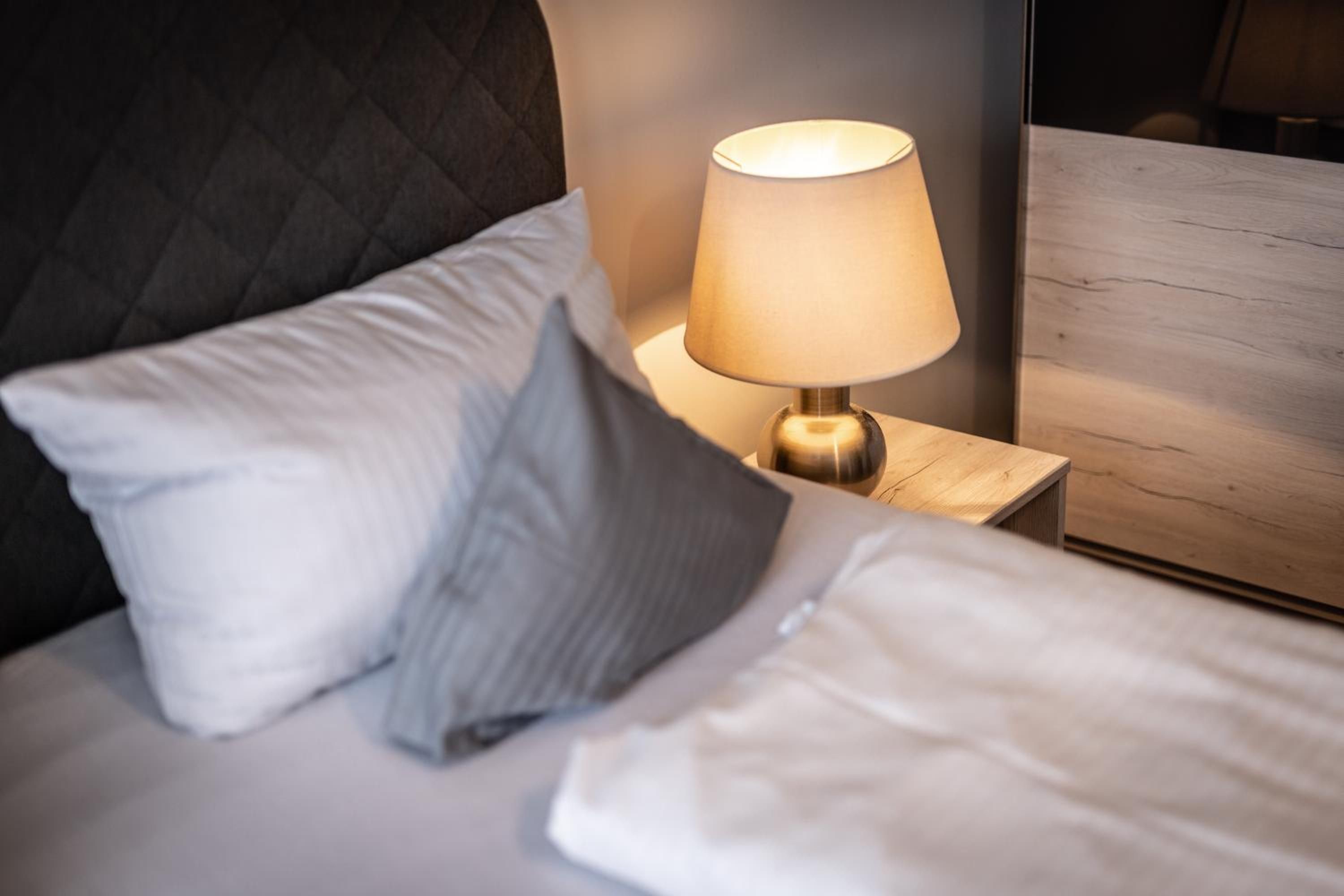 A neatly made bed with white bedding and two pillows, a bedside lamp on a wooden nightstand, and a dark headboard.