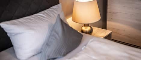 Una cama bien tendida con ropa de cama blanca y dos almohadas, una lámpara de noche sobre una mesita de madera, y un cabecero oscuro.