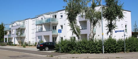 Un moderno edificio de apartamentos con balcones, una calle en su parte frontal y un coche estacionado al lado.