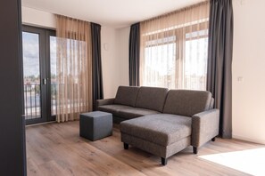 A modern living room with a sectional sofa, a small ottoman, and large windows with sheer curtains. - Living Neuburg (Neuburg)