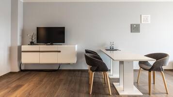 A modern living room with a flat-screen TV on a white cabinet, a dining table with four chairs, and two framed quotes on the wall.