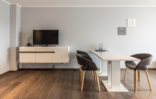 Uma sala de estar moderna com uma televisão de tela plana sobre um móvel branco, uma mesa de jantar com quatro cadeiras e duas frases emolduradas na parede.