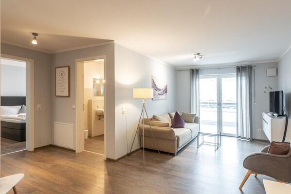 A modern hotel room with a bed, a sofa, a TV, and a view of the airport. - Living Neuburg (Neuburg)