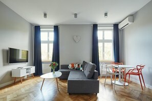 Un salon moderne avec un canapé gris, un téléviseur blanc, une petite table ronde et un coin repas avec des chaises rouges.
