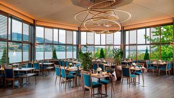 Un restaurant doté de nombreuses tables et chaises, de grandes fenêtres offrant une vue sur un plan d’eau et d’un luminaire moderne.