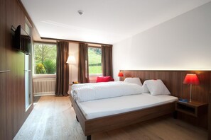 A hotel room with a large bed, wooden headboard, two bedside lamps, a TV, and a view of greenery through the window. - Seehotel Delphin (Meisterschwanden)