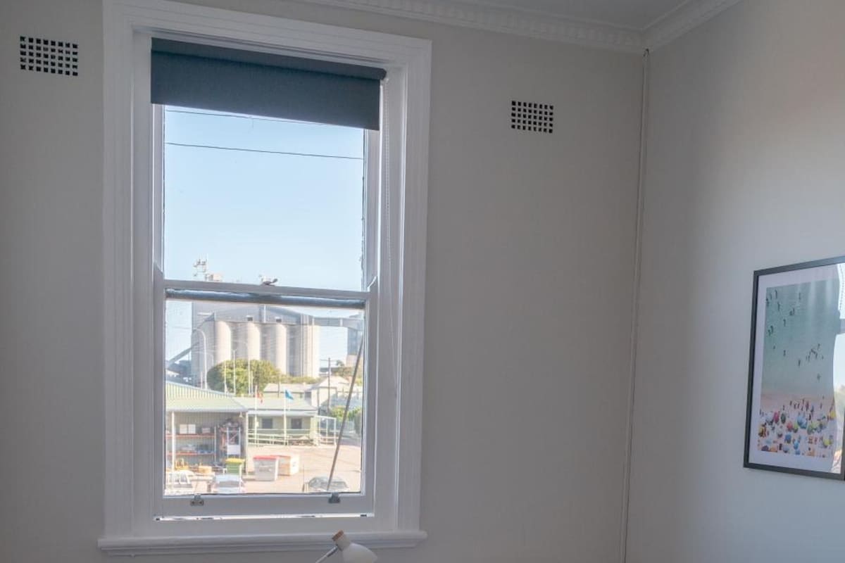A bedroom with a bed, a chair, a small table, and a window with a view of industrial buildings.