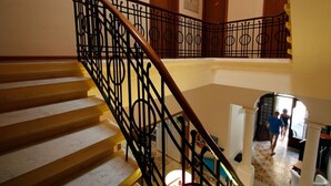A staircase with a wooden handrail and black metal balusters, leading to a landing with a door and a bulletin board. - Corner Hostel (Sliema)