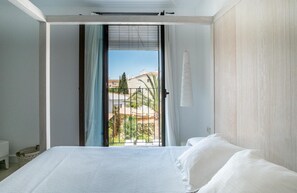 A bedroom with a bed, a window with a view of buildings and greenery, and a light-colored wall. - Hotel Es Marv S (Formentera)