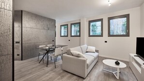 A modern living room with a sofa, a dining table, and chairs. There are windows with views of trees and a flat-screen TV on a stand. - La Perla By Sedar (Como)