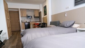 Quarto com duas camas, uma escrivaninha, uma cadeira, cozinha compacta e porta que dá para outro espaço.