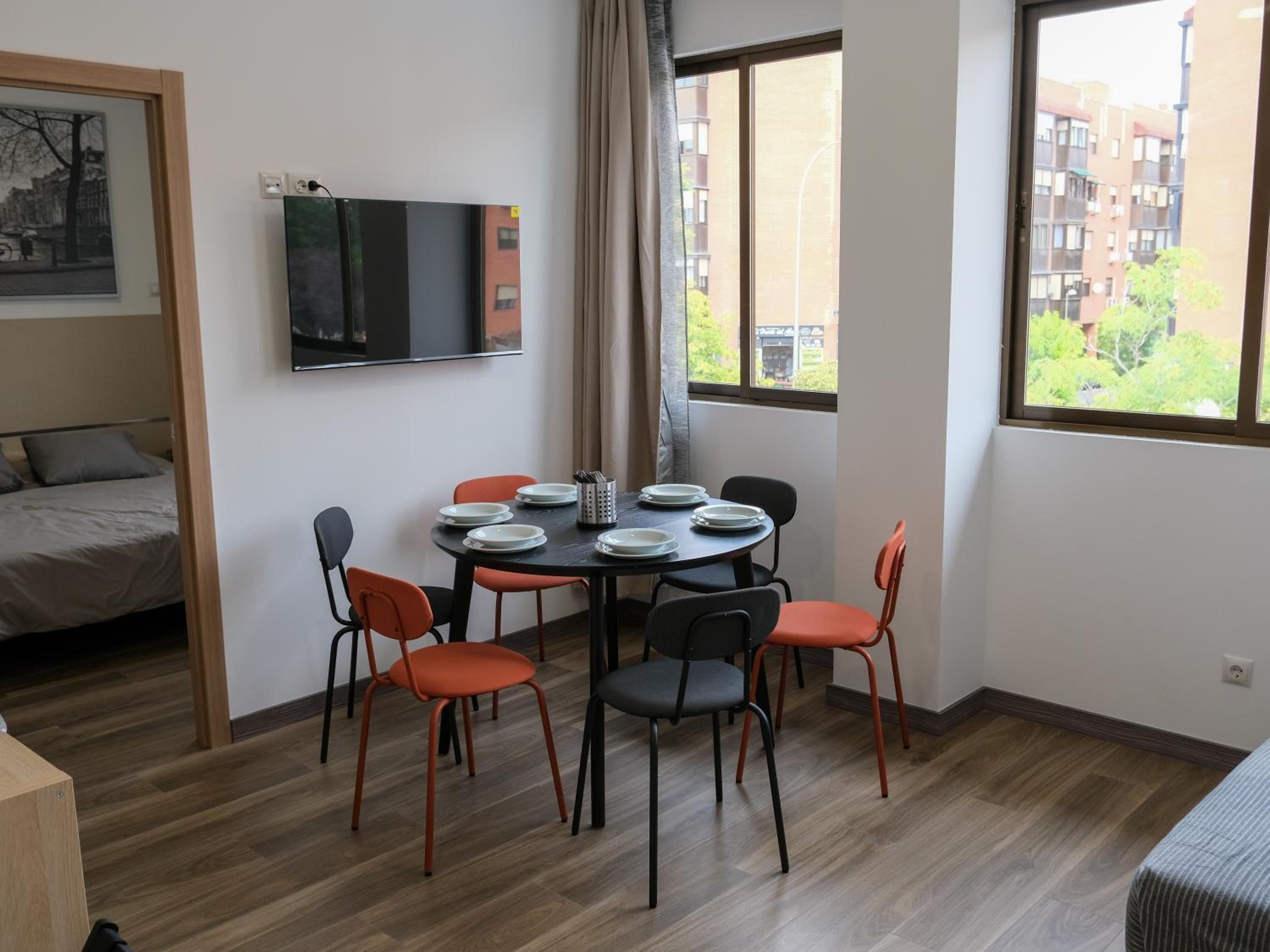 Una habitación con cama, una mesa de comedor con sillas, un televisor de pantalla plana montado en la pared y una ventana con vistas a edificios.