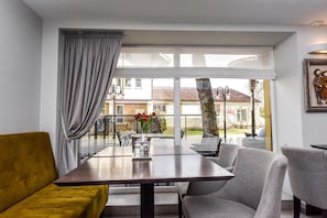 A modern living room with a sofa, a dining table, and chairs. There is a large window with curtains, and a view of the outside. - Muza Hotel (Palanga)