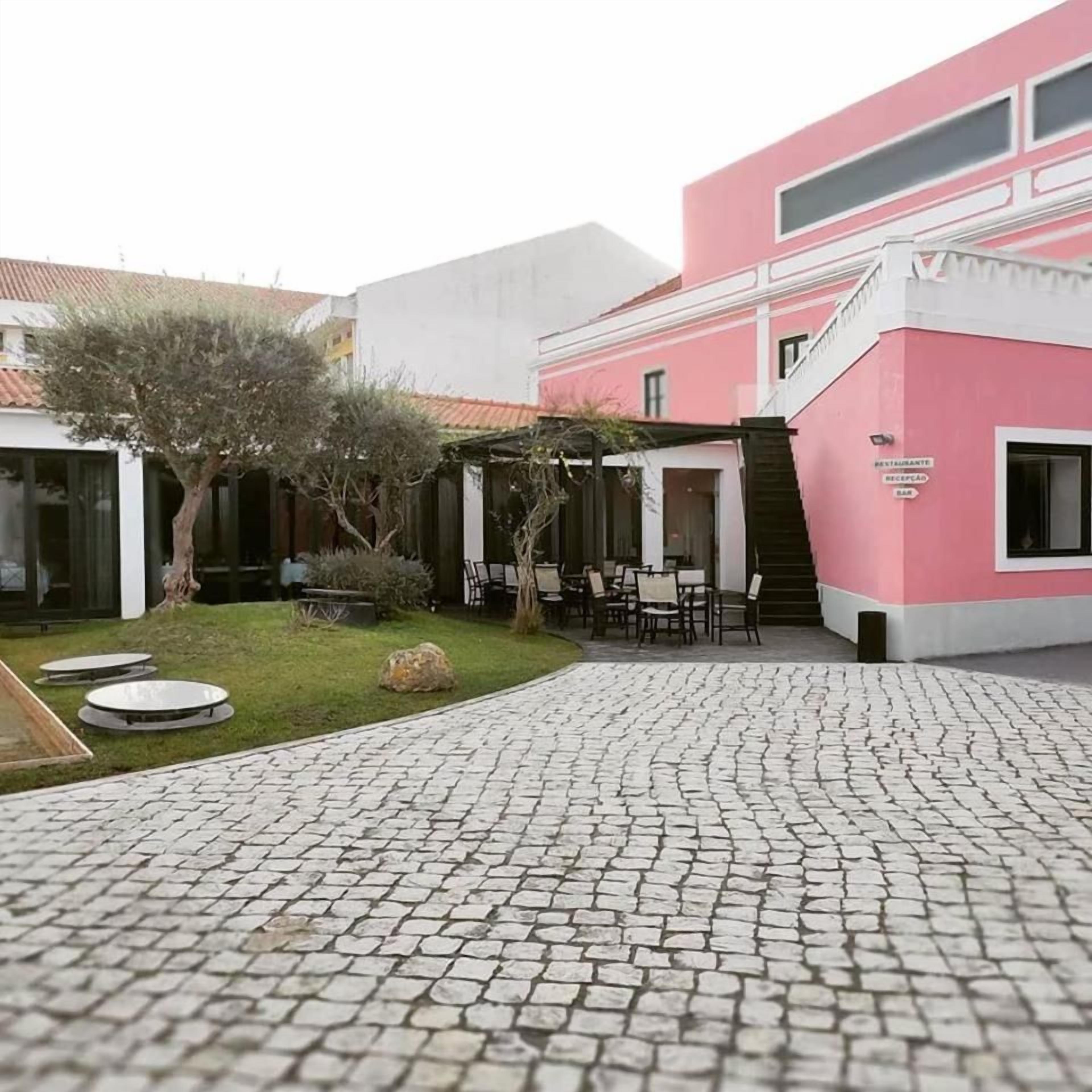 A courtyard with a cobblestone path, outdoor seating, and a building with a pink facade.