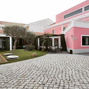A courtyard with a cobblestone path, outdoor seating, and a building with a pink facade. - Hotel Acez (Vendas Novas)