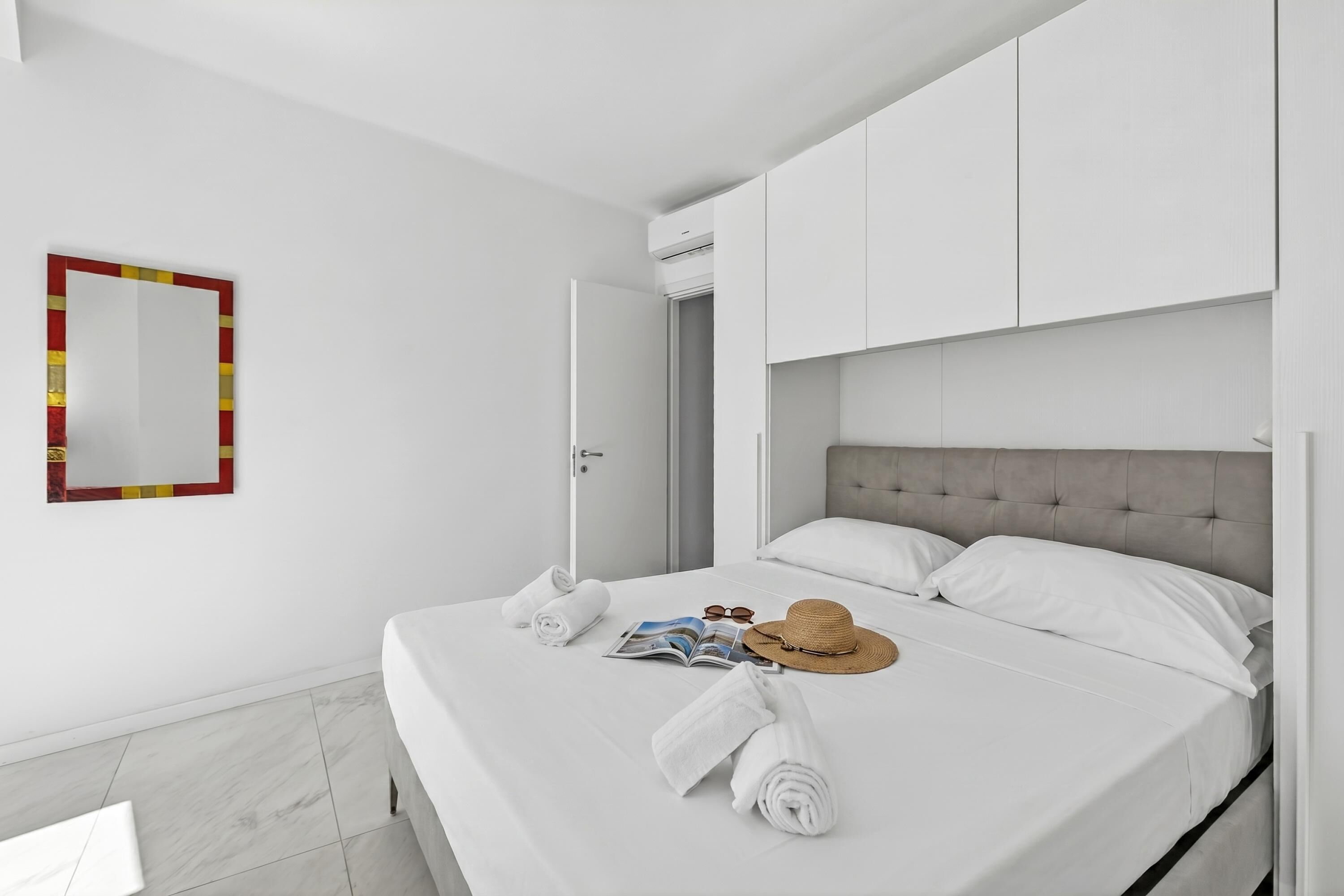 Cama arrumada com toalhas, um chapéu e óculos de sol, um espelho com moldura vermelha e uma porta que dá para outro quarto.