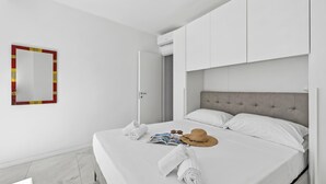 A neatly made bed with towels, a hat, and sunglasses, a mirror with a red frame, and a door leading to another room. - Emilia Sweet - with Balcony + AC, Sanremo, Italy (Sanremo)
