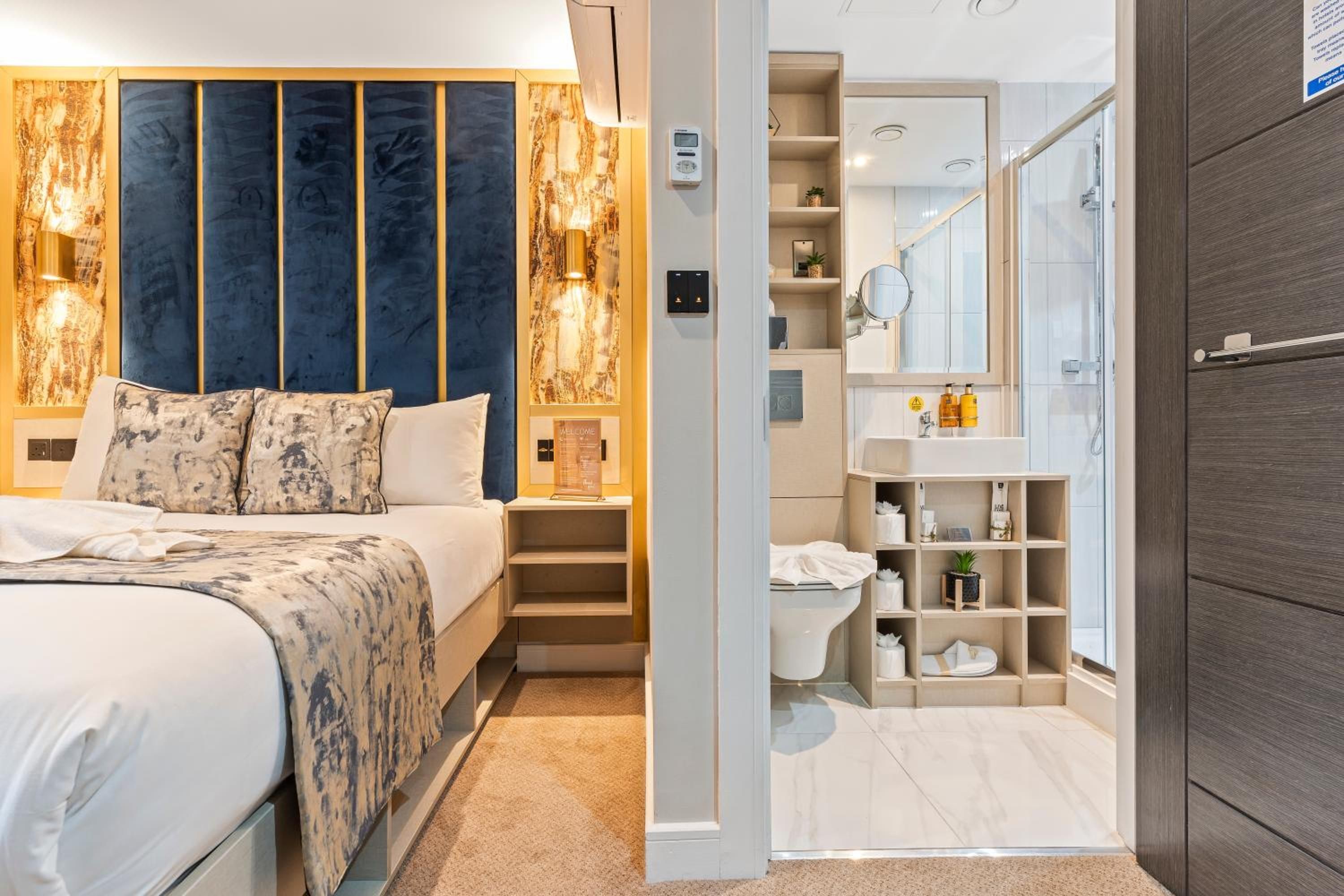 A modern hotel room with a large bed, a built-in headboard, a bedside table, a nightstand, a bathroom with a shower, a toilet, a sink, and a mirror.